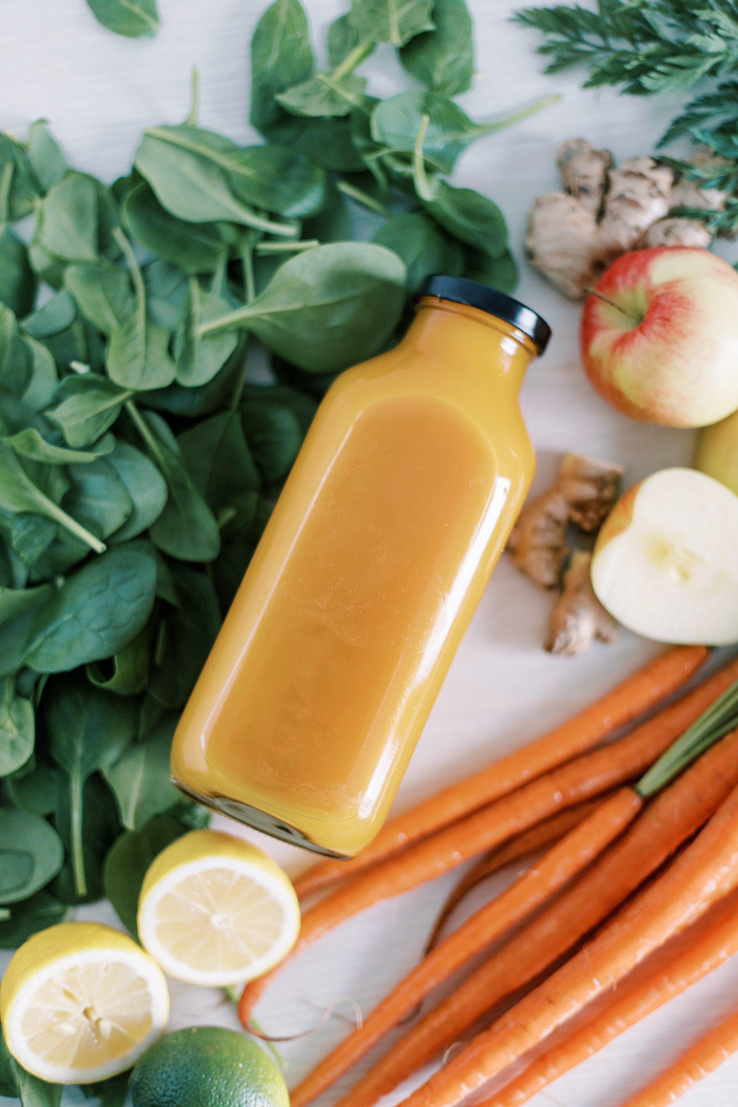 glass bottle of orange juice surrounded by carrots, lemons, spinach, ginger and apples