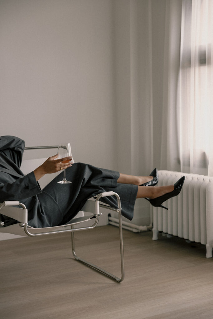 woman sat on chair wearing pant suit and heels holding a glass of red wine
